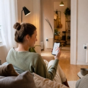 Smart Heizen in der Mietwohnung: Was ist erlaubt? – Person sitzt auf dem Sofa und steuert smarte Heizkörperthermostate per Smartphone-App – drei Thermostate sichtbar im Wohnzimmer einer Mietwohnung